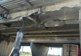 Image shows the underside of the Highway 17A overpass of the southbound side of Highway 99. Concrete and metal is torn away and hanging off the underside of the overpass. The oversized commercial container truck which struck the overpass is stopped and remains visible in the far right corner of the photo.